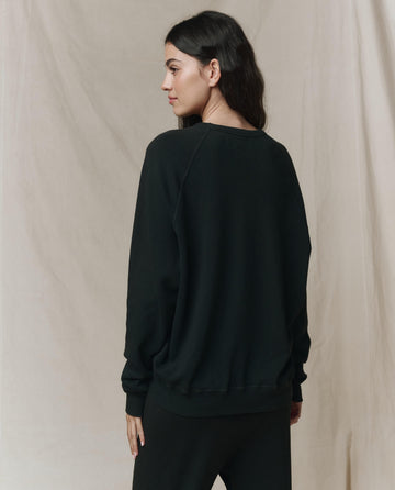 A woman with long dark hair faces away, wearing THE GREAT. CORE KNITS The College Sweatshirt. Solid -- Almost Black in 100% cotton and black pants, set against a neutral fabric backdrop.