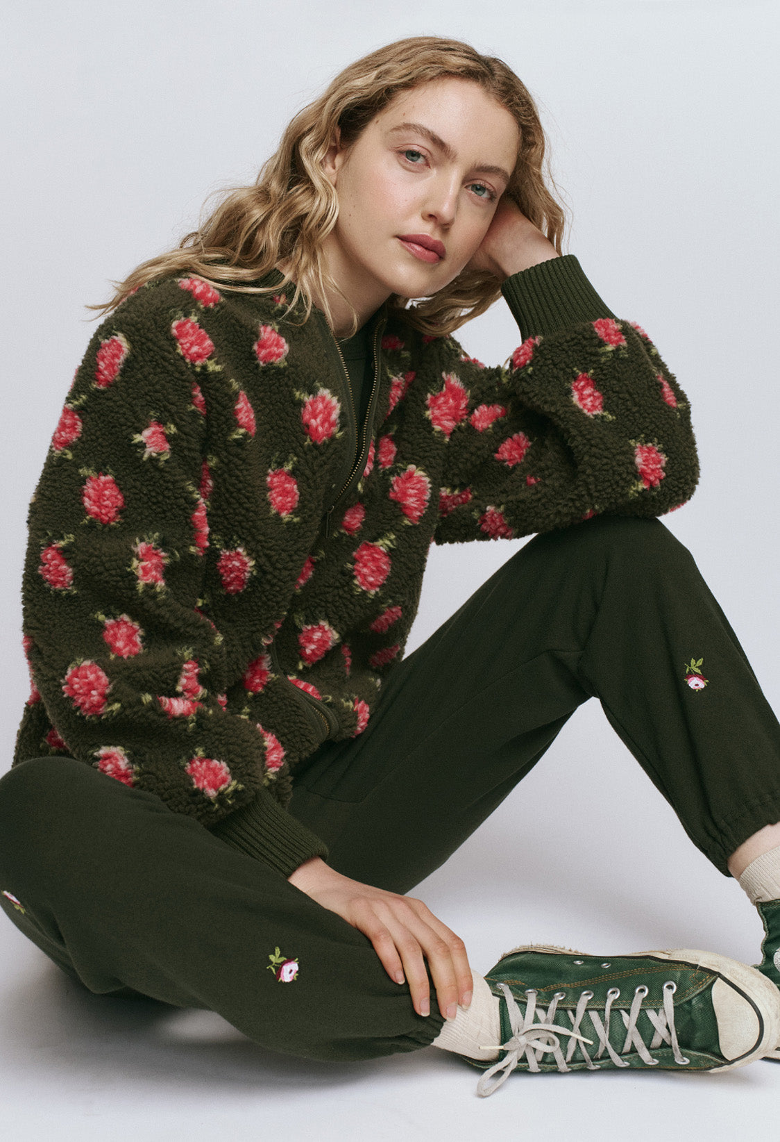 A woman with wavy blonde hair sits on the floor, wearing a dark green floral-patterned sweater, matching pants with small flower details, and green sneakers.