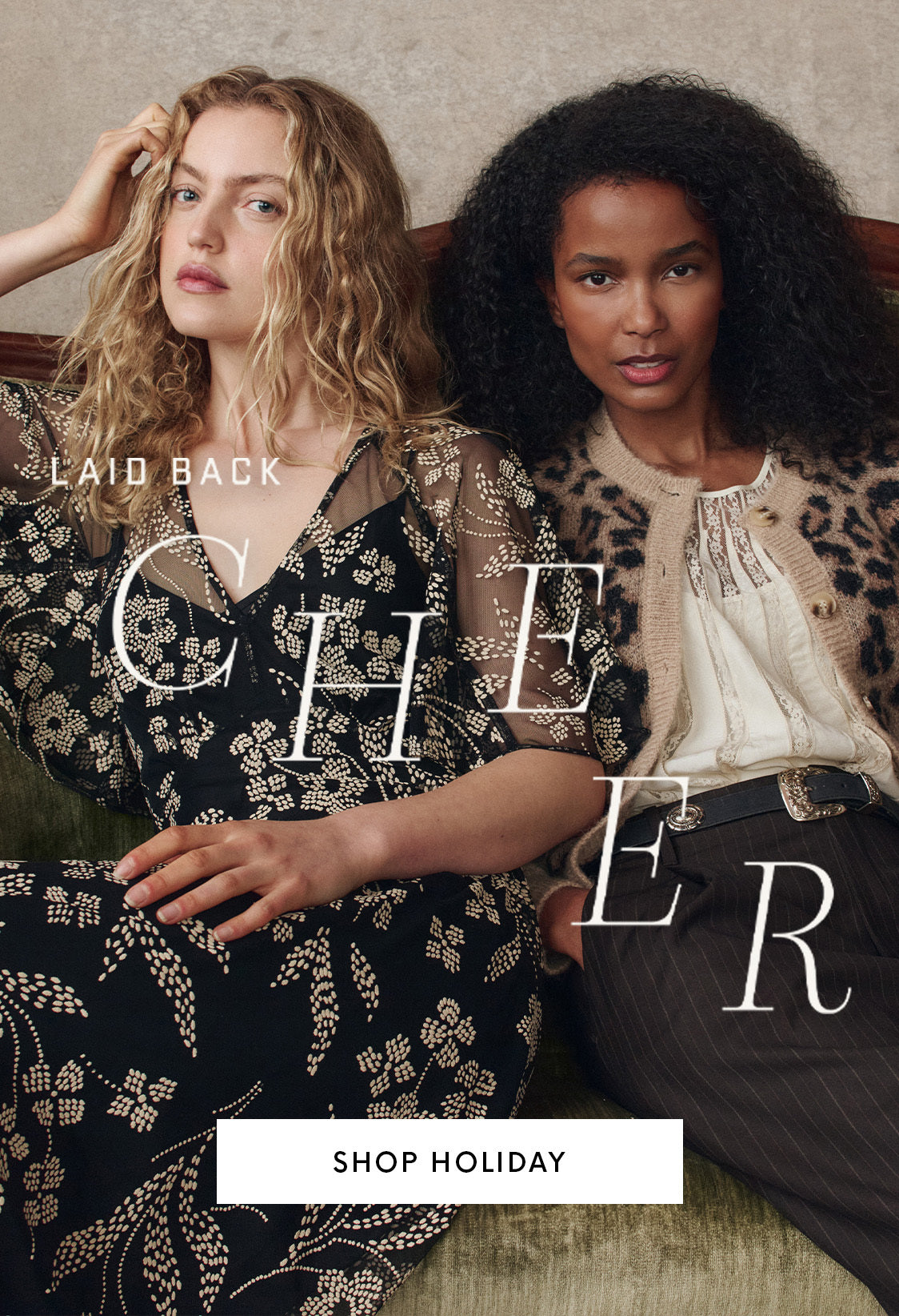 Two women sit on a green couch wearing patterned holiday outfits, one in a black dress with gold details, the other in a white top and leopard print cardigan. Text reads "Laid Back Cheer. Shop Holiday.