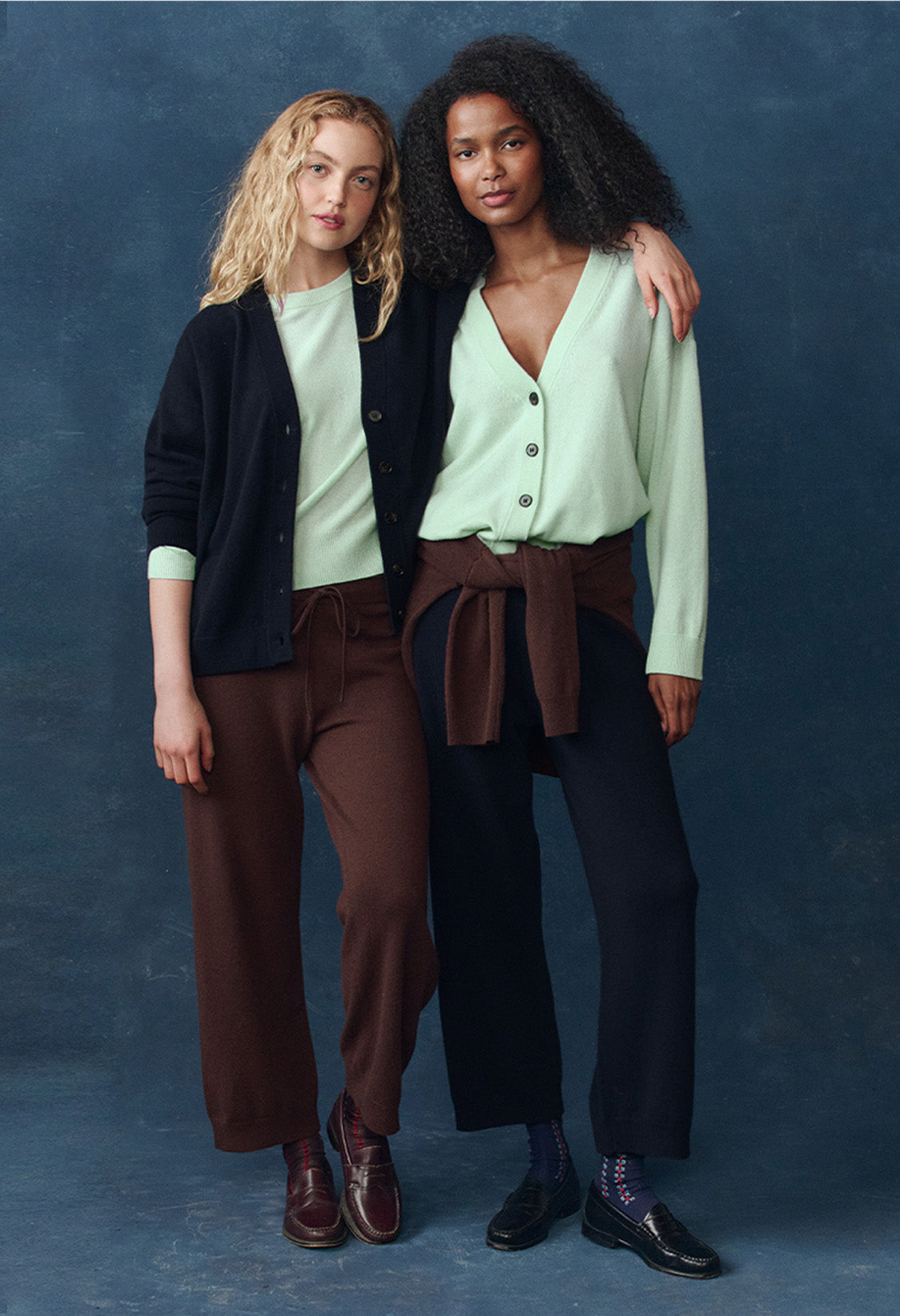 Two women stand side by side with arms around each other, wearing cardigans, sweaters, loose pants, and loafers against a plain blue background.
