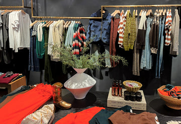 A retail display showing assorted clothing on hangers, folded garments on a table, a large white vase with greenery, a pair of golden boots, and stacked boxes of products.