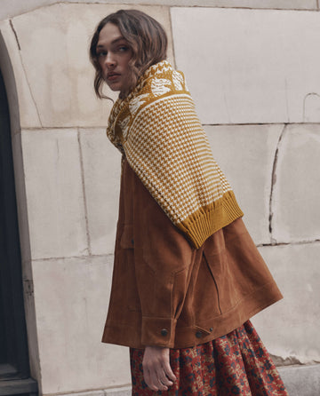 Wearing THE GREAT.’s The Suede Folsom Jacket in Cognac, a person with a patterned scarf and floral skirt stands in front of a textured stone wall, glancing over their shoulder.