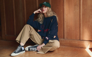 Person with long curly hair wearing a green cap, navy sweater, khaki pants, and black sneakers sits on a wooden floor against a paneled wall.