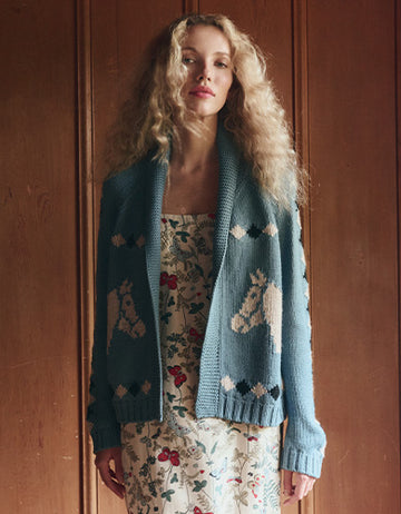 A woman with curly blonde hair wears a floral dress and a blue cardigan featuring horse head patterns, standing in front of a wooden wall.