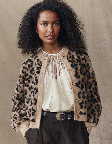 Woman standing against a neutral background wearing a leopard print cardigan, a white lace top, dark pinstripe pants, and a black belt with a decorative buckle.