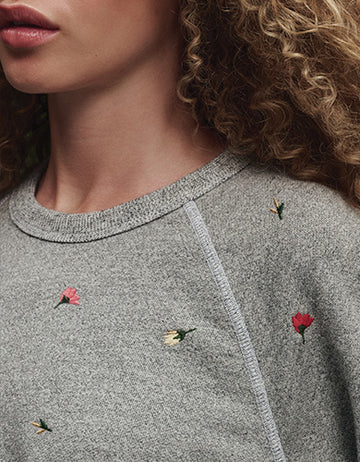 Close-up of a person wearing a grey sweater with small embroidered flowers. Only part of their face, neck, and curly hair are visible.