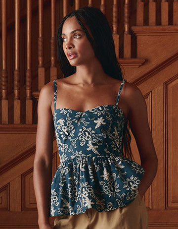 A woman with long hair stands indoors in front of wooden stairs, wearing a blue and white floral-patterned peplum top and beige pants.