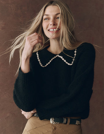 Woman with long blonde hair wearing a black sweater with a white scalloped collar and tan pants, standing against a brown background and smiling.