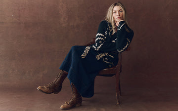A woman sits on a chair against a brown background, wearing a long patterned sweater, a long skirt, and lace-up brown boots, with her chin resting on her hand.