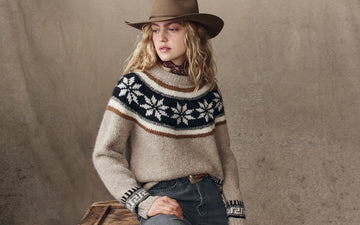 A person wearing a brown wide-brim hat, a patterned knit sweater with snowflakes, jeans, and a brown belt sits against a brown textured background.
