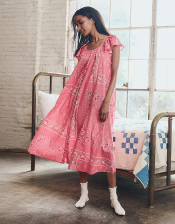 A woman stands indoors beside a metal bed, wearing a loose pink dress with a bandana pattern and white socks.