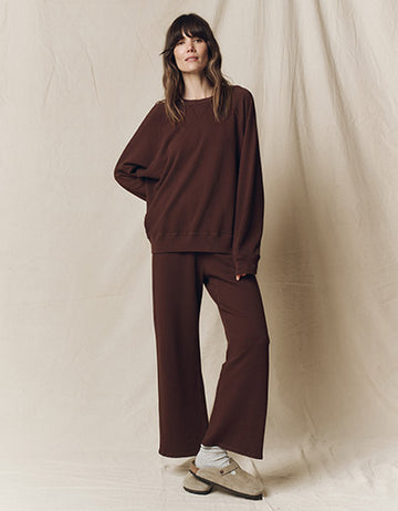 A woman stands against a beige fabric backdrop, wearing a loose-fitting brown sweatshirt and matching wide-leg pants with light-colored shoes and socks.