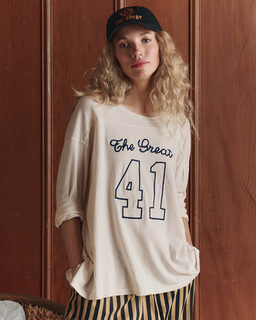 A person wearing a black cap, an oversized cream shirt with "The Great 41" printed on it, and striped pants stands in front of a wooden wall.