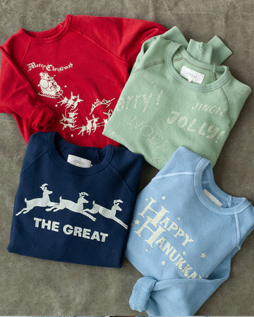 Four folded holiday-themed sweatshirts on a gray surface, featuring Christmas, Hanukkah, and festive designs in red, green, navy, and light blue.