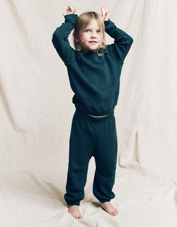 Young child standing barefoot on fabric backdrop, wearing a dark green long-sleeve top and matching pants, with arms raised above their head.