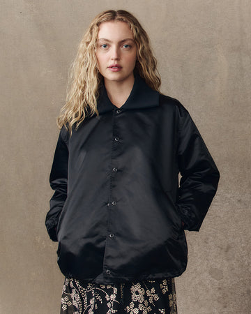 A woman with wavy blonde hair wears a black button-up jacket and patterned pants, standing against a plain, textured background.