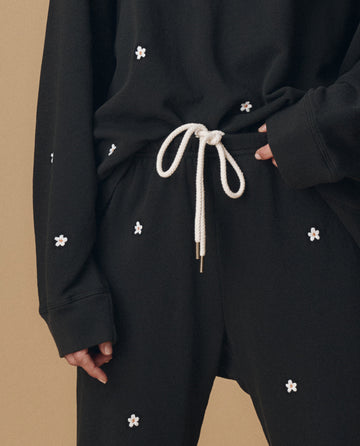 A person wears THE GREAT. PF25 EMB KNITS' The Stadium Sweatpant in Almost Black with Daisy Embroidery, featuring a white drawstring, against a beige background.