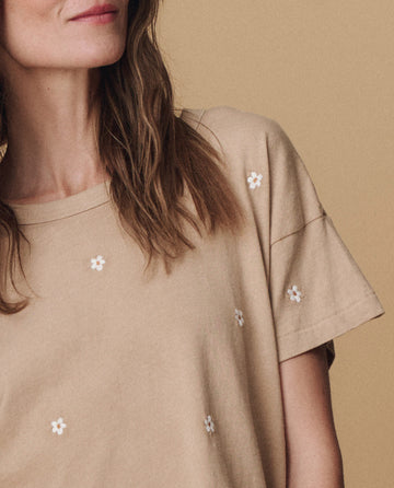 A woman wears THE GREAT. PF25 EMB KNITS 'The Boxy Crew. Embroidered' t-shirt in khaki with daisy embroidery, standing against a matching background. Only her lower face, neck, and upper torso are visible.