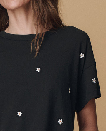 A woman is shown from the shoulders up against a beige background, wearing THE GREAT. PF25 EMB KNITS The Boxy Crew in Almost Black with daisy embroidery.