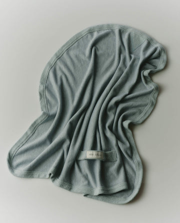 The RŌZ + The GREAT The Sweatshirt Hair Towel in Seaglass by THE GREAT. PF25 ROZ is a soft, light blue towel with a wavy edge and sewn label, designed for quick drying and frizz reduction, shown spread flat on a surface.