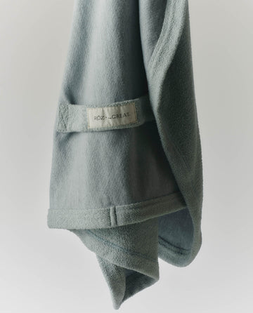 A light green RŌZ + The GREAT The Sweatshirt Hair Towel in Seaglass, from THE GREAT. PF25 ROZ, hangs against a plain white background—ideal for quick drying and reducing frizz.