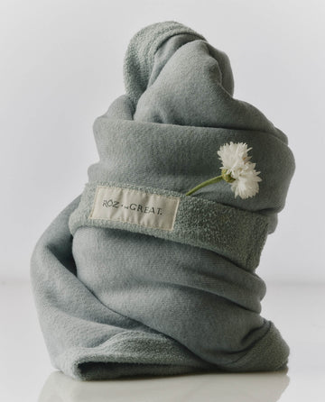 A Seaglass green RŌZ + The GREAT The Sweatshirt Hair Towel by THE GREAT. PF25 ROZ is rolled upright with small white flowers under the label—designed to reduce frizz and dry hair quickly.