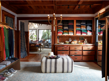 Clothing boutique interior with folded clothes on shelves, a central ottoman, wooden drawers, and a chandelier, leading into a room with a table and window view of trees.