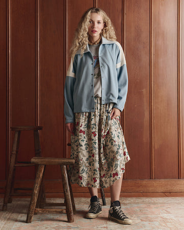 A woman stands indoors by wooden paneling, wearing a blue jacket, graphic t-shirt, floral skirt, and black high-top sneakers. A wooden stool is positioned beside her.