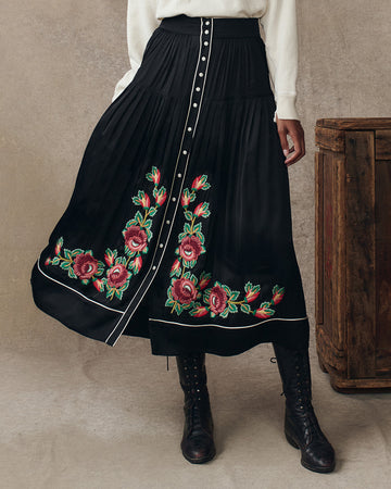 A person wearing a black pleated skirt with red floral embroidery, a white top, and black lace-up boots stands next to a wooden chest.