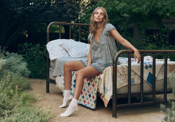 A woman in patterned pajamas and socks sits on the edge of a metal bed outdoors, surrounded by quilts and greenery.