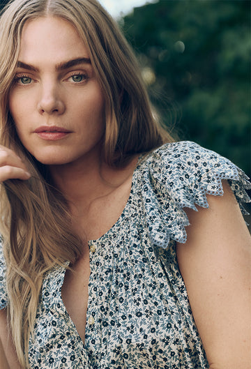 Woman with long blonde hair wearing a floral blouse with lace-trimmed sleeves, looking at the camera against a blurred outdoor background.