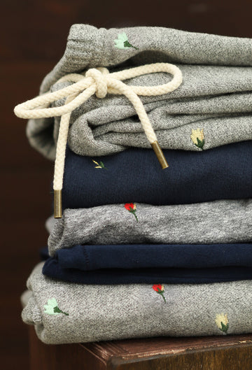 A neatly folded stack of sweatpants in gray and navy, each with small embroidered flower designs and visible white drawstrings on the top pair.