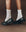 Someone wearing THE GREAT. F25 D1 ACCESSORIES' The Monogrammed Trouser Sock in Cream, featuring "TG" monogramming, and black loafers, stands on a brown surface.