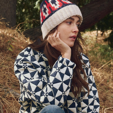 A woman wearing a patterned jacket and a knit hat sits outdoors on dry grass, resting her chin on her hand and looking to the side.