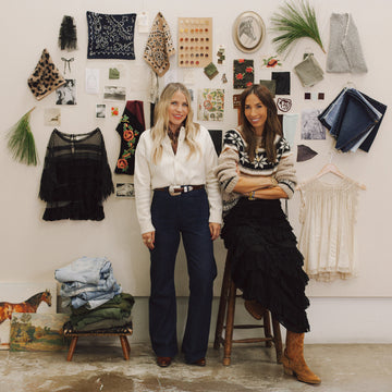 Two women stand in front of a wall with fabric swatches, art, and notes. One is standing, the other is seated on a stool. Folded clothes and a horse picture are on the floor nearby.