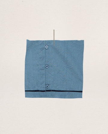 The Western Sleep Cami in Blue Dusk by THE GREAT. SP25 SLEEP, shown as a square blue fabric with three vertical buttons and a thin pin at the top, is set against a plain off-white background.