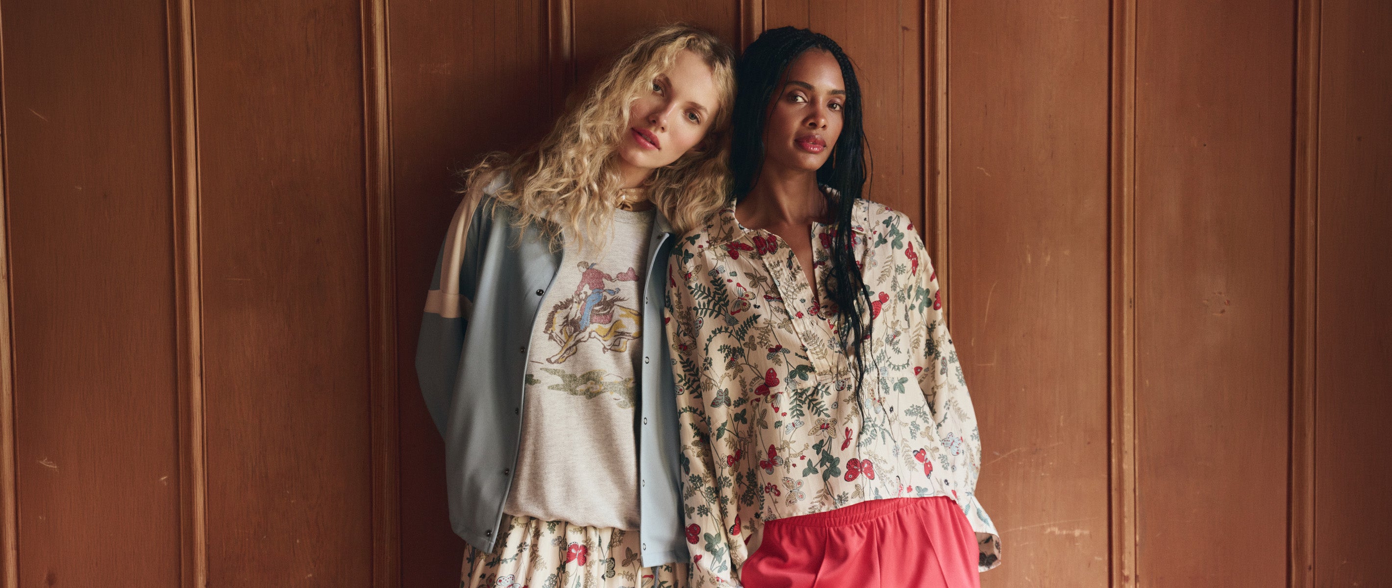 Two women stand against a wooden wall; one wears a graphic t-shirt and skirt, the other wears a floral blouse and red skirt.