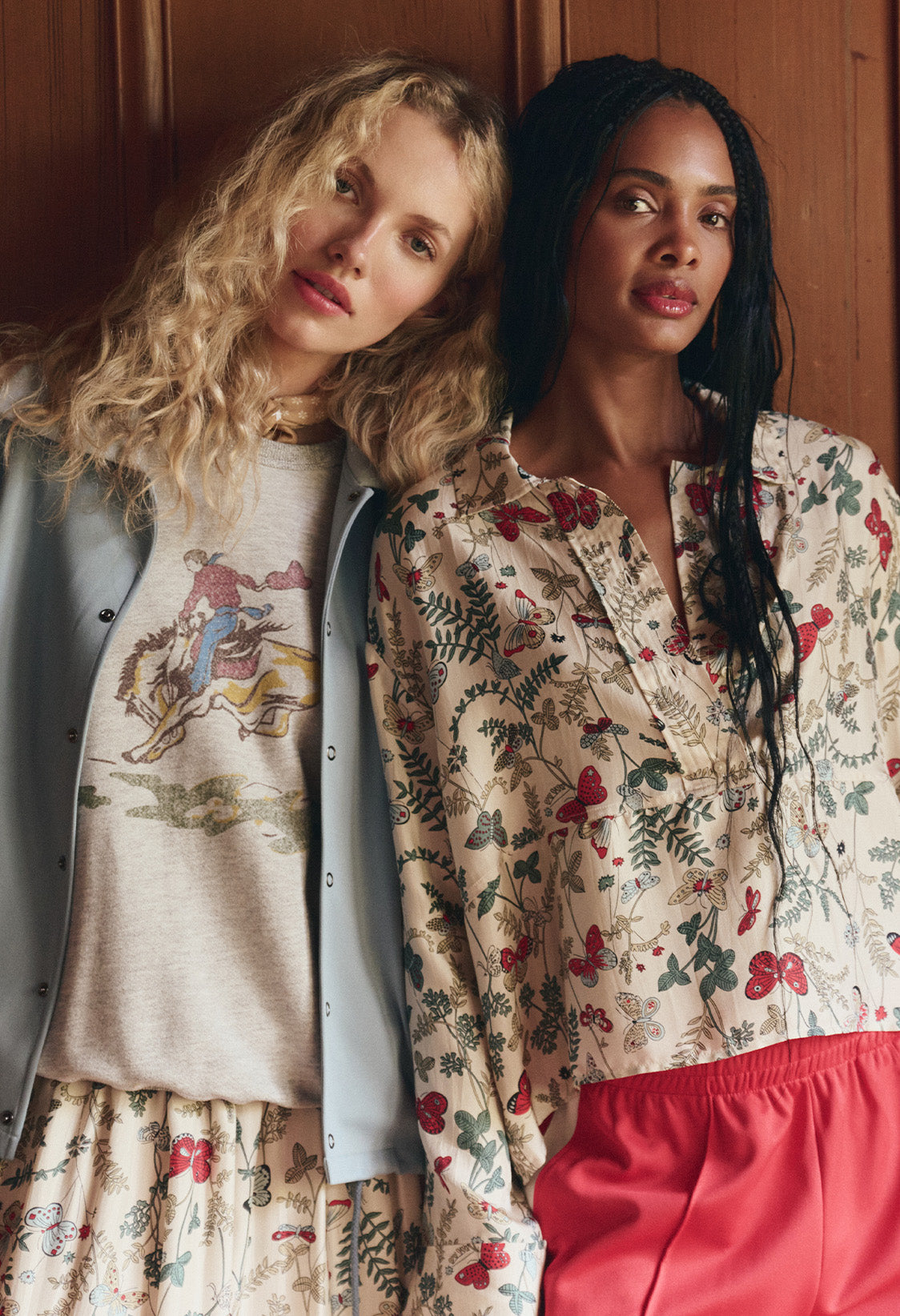 Two women standing side by side, one wearing a graphic sweatshirt with a blue vest and skirt, the other in a floral-patterned blouse with red pants, both looking at the camera.