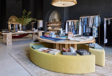 A boutique clothing store interior with folded clothes on a round wooden display table, racks of hanging garments, and modern decor.