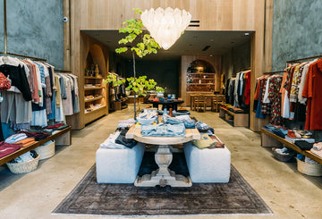 Boutique clothing store interior with racks of colorful clothes, wooden shelves, central table displaying folded garments, and a chandelier overhead.