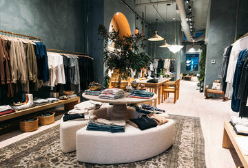 Clothing store interior with racks of neatly arranged clothes, a central round display table with folded garments, wooden counters, potted plants, and ambient lighting.