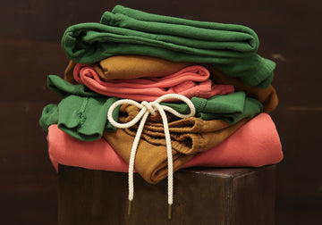 A neatly folded stack of green, brown, and coral-colored clothing, with a pair of pants featuring a white drawstring, placed on a dark wooden surface.