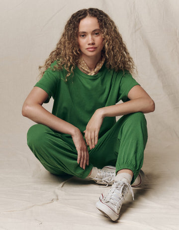 Person with curly hair sits on the floor wearing a green t-shirt, matching green pants, white sneakers, and a light-colored scarf, against a neutral backdrop.