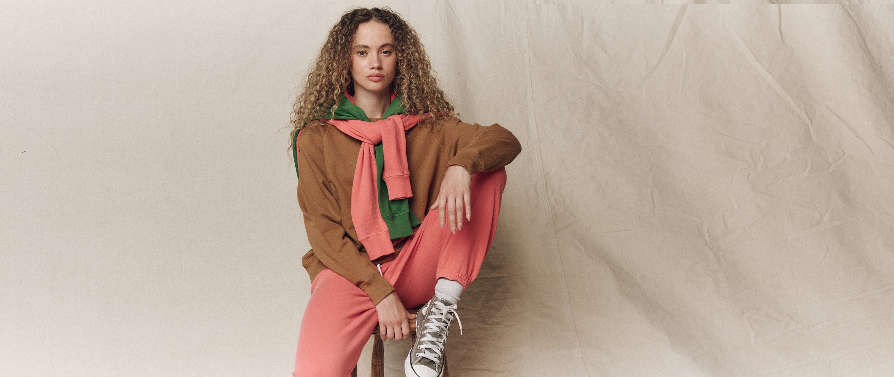 A person with curly hair sits on a stool wearing a brown jacket, green hoodie, pink sweatshirt, pink pants, and sneakers, posed against a beige fabric backdrop.