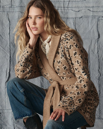 A woman with long, wavy blonde hair wearing a tan floral belted cardigan, blue jeans, and a white scarf sits on a stool against a textured gray backdrop.