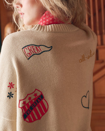 A person with wavy blonde hair wears a beige sweater featuring embroidered patches, including a shield, a heart, and words like “GREAT” and “all-star,” with a red patterned collar underneath.