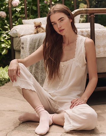 A woman in a white nightgown and pink socks sits on a stone floor near a bed outdoors, with greenery and a teddy bear in the background.