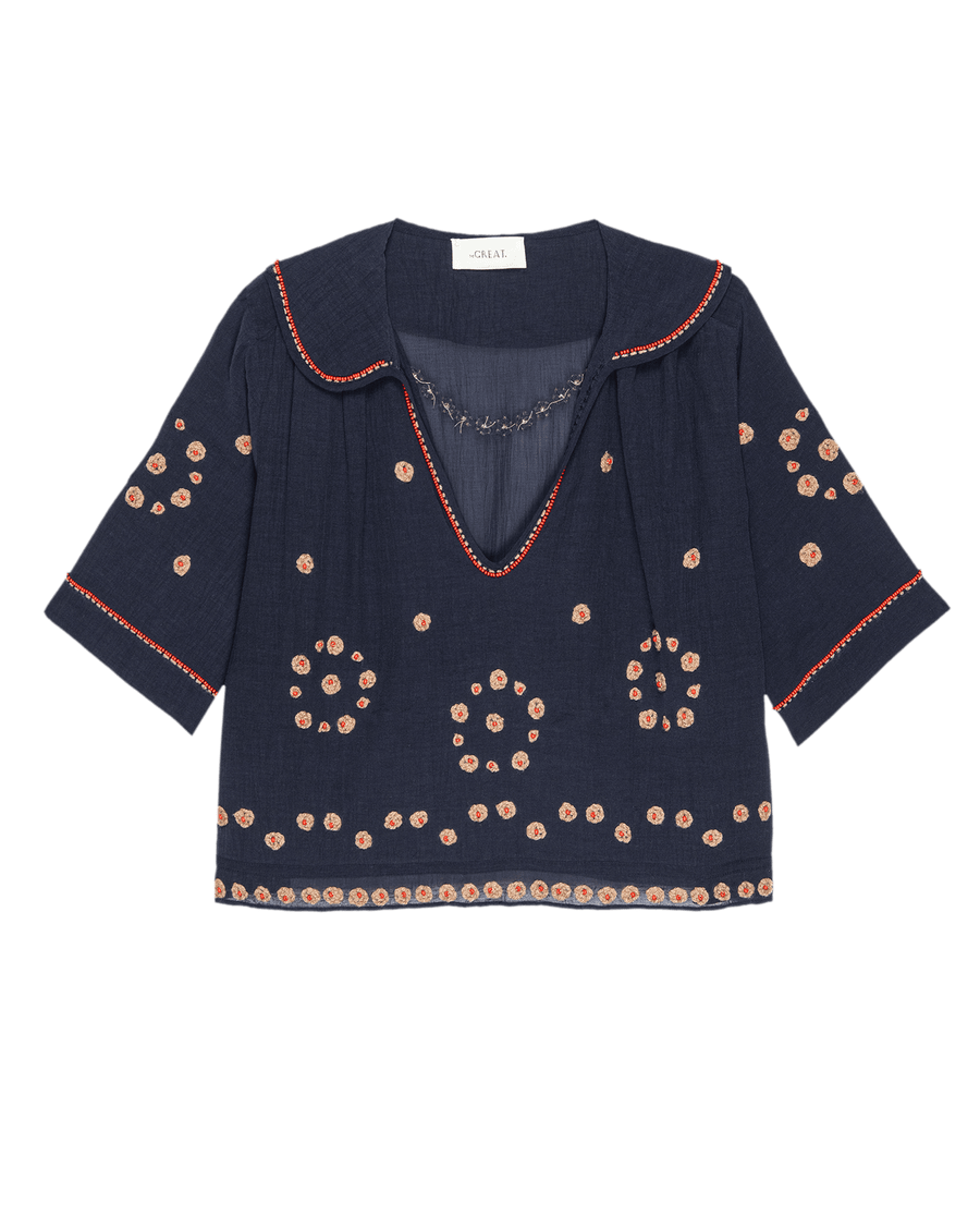 The Beaded Vintage Sailor Top by THE GREAT. SU25 is a navy blue sailor-style top with half sleeves, red embroidered edges, and beige and red circular floral patterns throughout the fabric.
