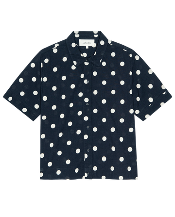 The Messenger Top by THE GREAT. PS26 features a navy background with cream polka dots, short sleeves, and a boxy fit in lightweight cotton, shown laid flat on a white surface.