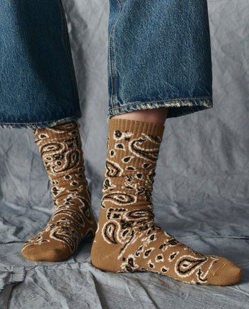 Someone wearing THE GREAT. H25 COLLECTION The Bandana Sock in Camel stands on a gray fabric background, paired with cropped blue jeans.